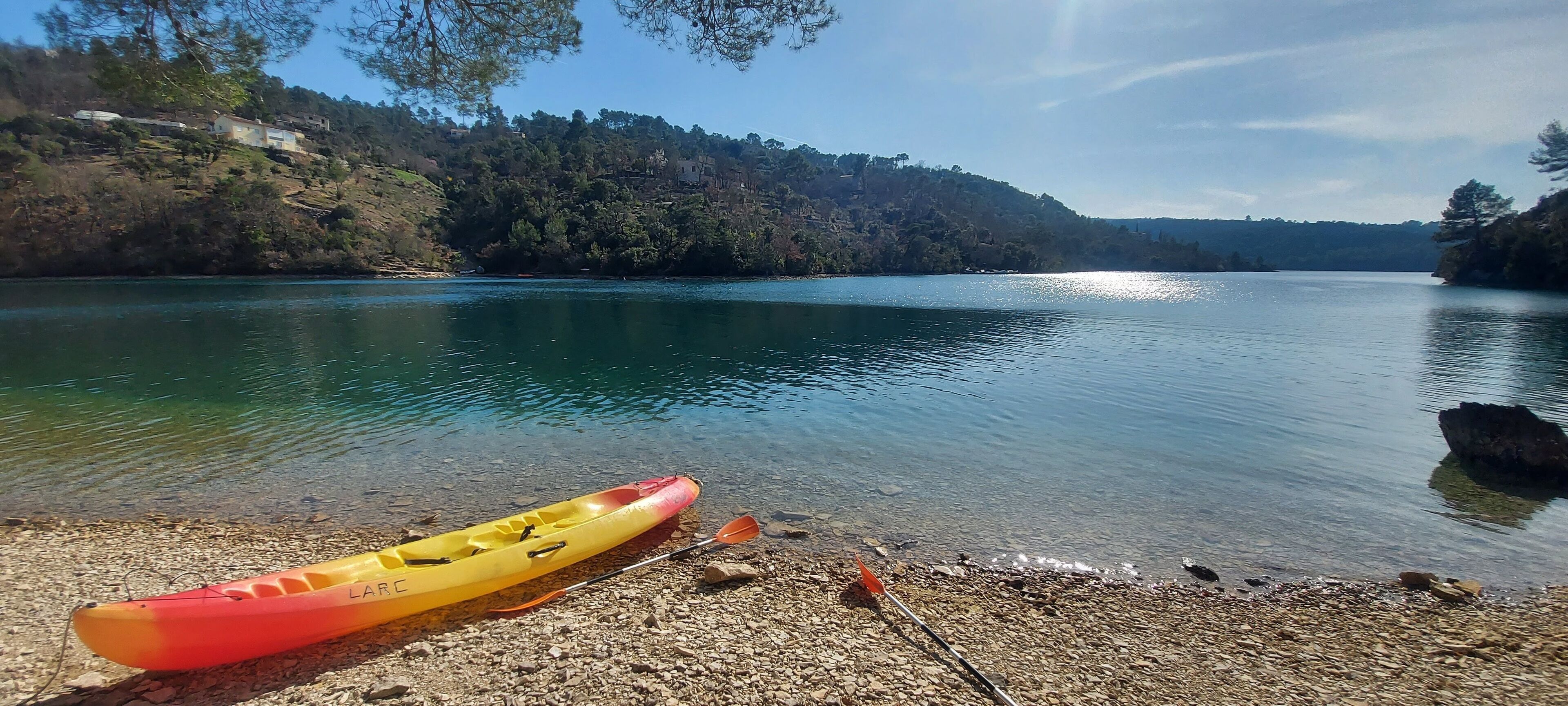Un kayak sobre una orilla pedregosa junto a un lago tranquilo, con casas en una colina al fondo.