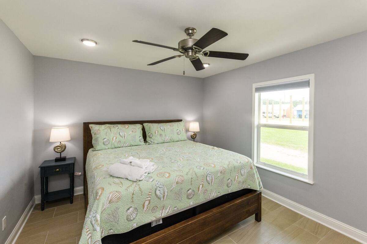 A bedroom with a bed, two bedside tables, a ceiling fan, and a window with a view of a grassy area.