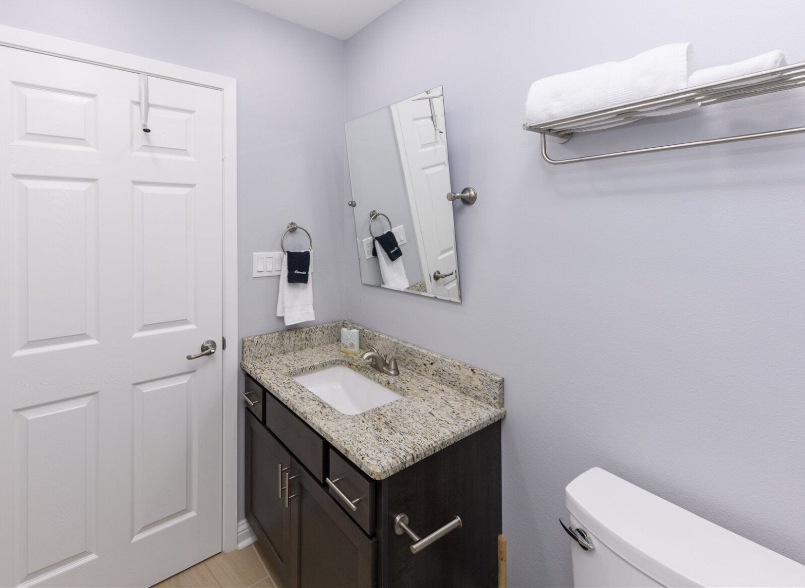 Un baño con puerta blanca, un lavabo con encimera de granito, un mueble oscuro, un inodoro, un perchero y un espejo.