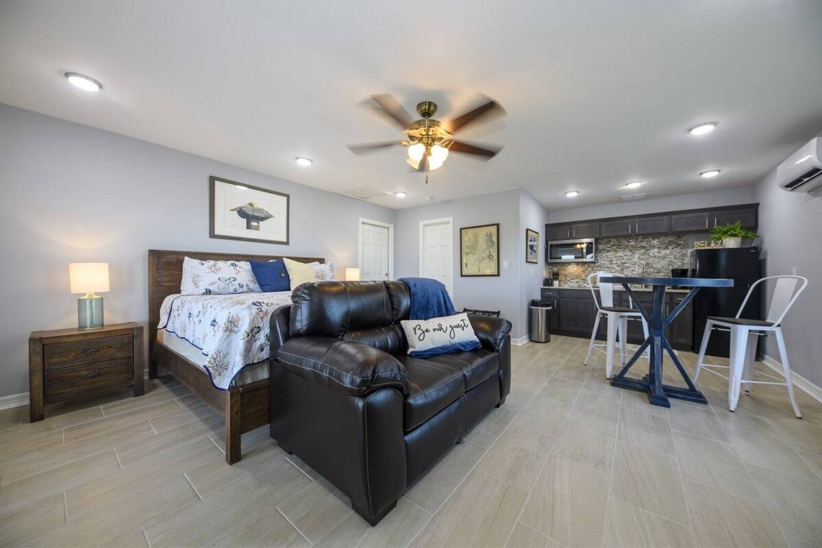 A modern living room with a leather sofa, a dining table with chairs, a kitchen area, and a ceiling fan.