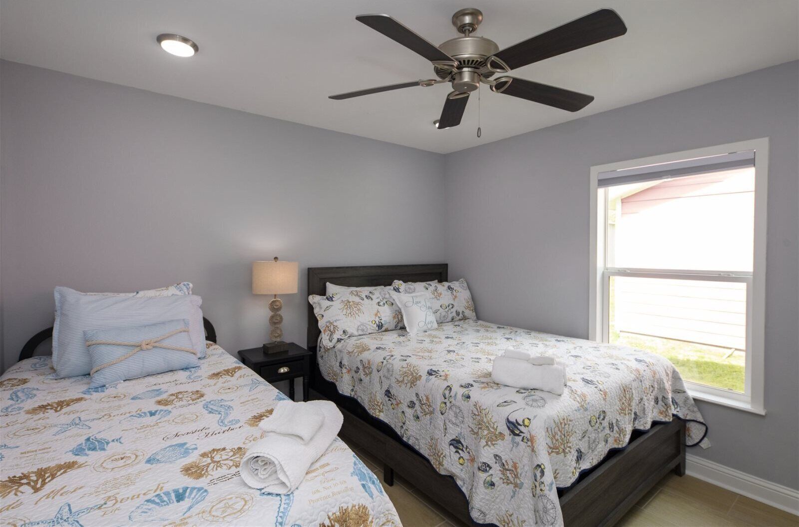 A bedroom with two beds, a ceiling fan, a window, and a nightstand with a lamp.