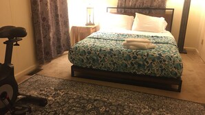 A bedroom with a bed, a nightstand, a lamp, and a stationary exercise bike.