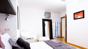 Quarto de hotel moderno com uma cama grande, uma escrivaninha e uma cadeira. Há uma TV fixada na parede e uma porta que dá para uma varanda.