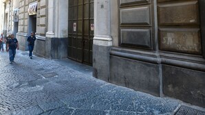 Fachada de um edifício histórico com grandes portas e janelas, rua de paralelepípedos e alguns pedestres.