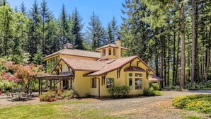Una casa de dos pisos con fachada amarilla y techo de tejas rojas, rodeada por un bosque.