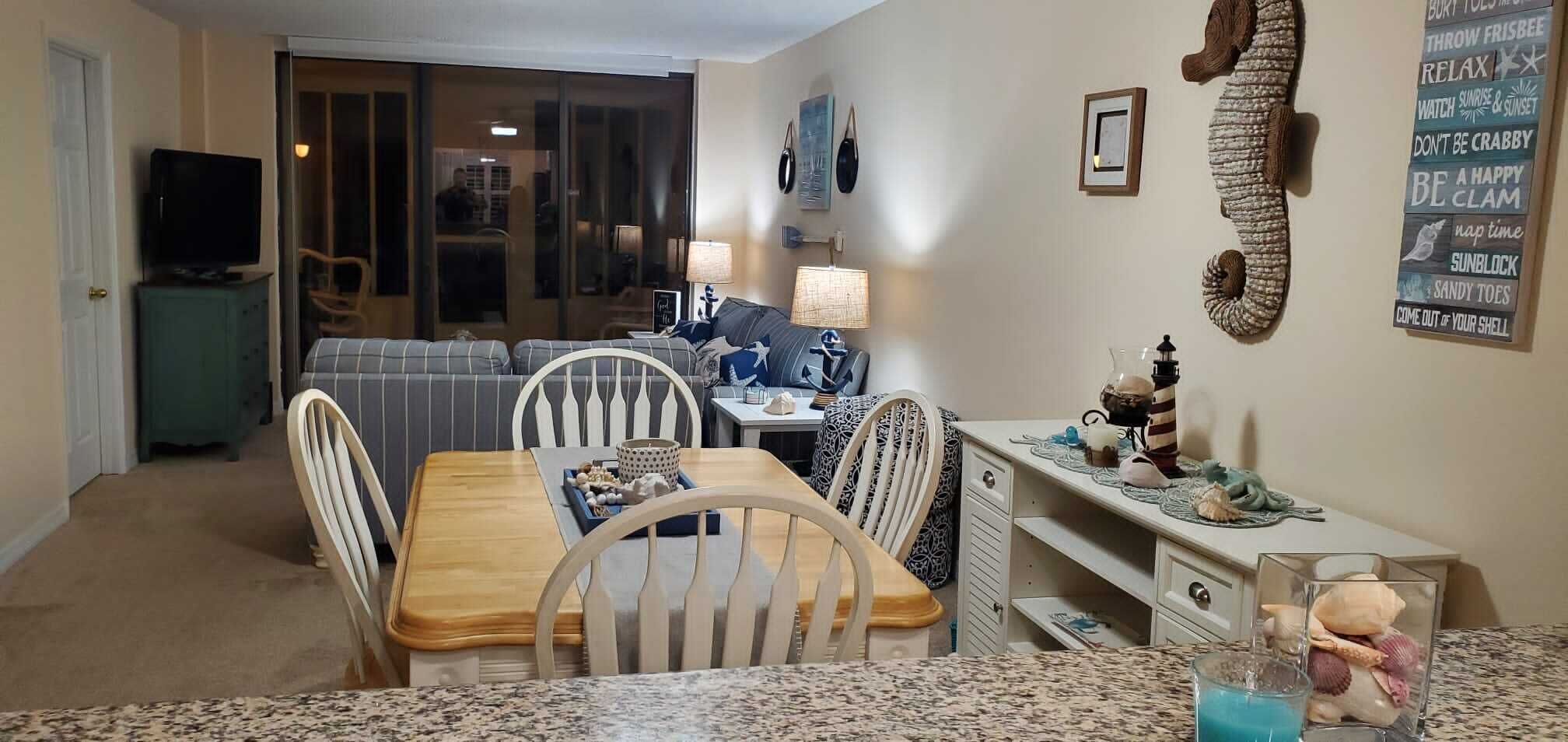 A dining area with a wooden table and chairs, a kitchen counter with decorative items, and a living area with a sofa and lamps.