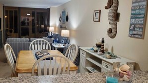 A dining area with a wooden table and chairs, a kitchen counter with decorative items, and a living area with a sofa and lamps.