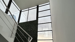 A modern staircase with metal railings and a glass railing, leading to a landing with a view of the cityscape through large windows. - 3BR 10 Guest with Pvt. Jacuzzi 2 pools Gated Comm. AC Security 24-7 (Rosarito)