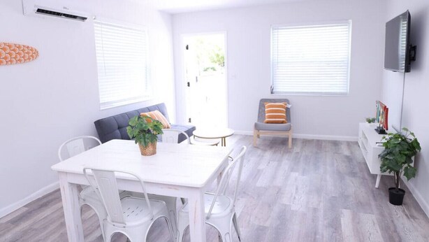 A modern living room with a white table and chairs, a grey sofa, a TV mounted on the wall, and a large window with blinds. - 1111 Douglas · Remodeled 1bed/1bath apt w/parking great location (Miami)