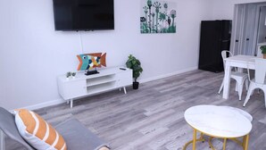 A modern living room with a flat-screen TV, a white entertainment unit, a colorful fish decoration, a small potted plant, a grey sofa with an orange cushion, and a white dining table with chairs. - 1111 Douglas · Remodeled 1bed/1bath apt w/parking great location (Miami)