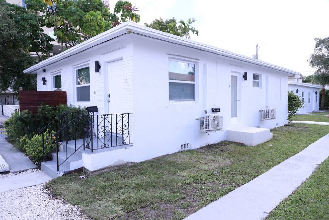 A single-story white house with a front porch, two windows, and an air conditioning unit.
