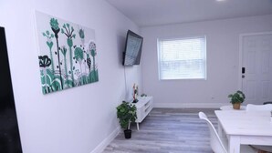 Ein geräumiges Zimmer mit weißer Wand, einem Wand-Fernseher, einem Fenster mit Jalousien, einem weißen Esstisch mit Stühlen und einem Topf mit Pflanze.