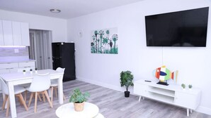 Ein modernes Wohnzimmer mit einem weißen Sofa, einem Esstisch mit Stühlen, einem kleinen runden Tisch mit einer Pflanze, einer weißen Unterhaltungsecke mit einem Fernseher und einem Gemälde an der Wand.