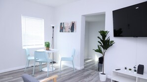 A modern living room with a grey sofa, a round dining table with two chairs, a large potted plant, a flat-screen TV mounted on the wall, and a white console table with a few items on it. - 1113 Douglas rd · Cozy Studio w/ parking & private patio w/ BBQ (Miami)