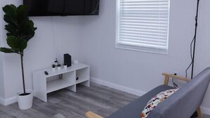 A modern living room with a grey sofa, a mounted TV, a white entertainment unit, a potted plant, and a window with blinds. - 1113 Douglas rd · Cozy Studio w/ parking & private patio w/ BBQ (Miami)