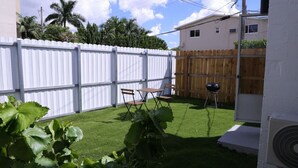 A backyard with artificial grass, a wooden table and chairs, a barbecue grill, and a white fence. - 1113 Douglas rd · Cozy Studio w/ parking & private patio w/ BBQ (Miami)