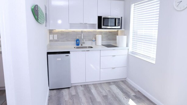 A compact kitchen with white cabinets, a microwave, a dishwasher, and a window with blinds. - 1113 Douglas rd · Cozy Studio w/ parking & private patio w/ BBQ (Miami)