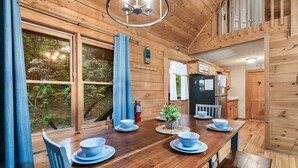 Interior de uma cabana de madeira com uma mesa de jantar posta para quatro pessoas, um pequeno arranjo de plantas no centro e uma área de cozinha com geladeira e armários.