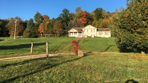 Une maison à deux étages avec un porche à l’avant, entourée d’un champ verdoyant et d’arbres aux feuilles d’automne.