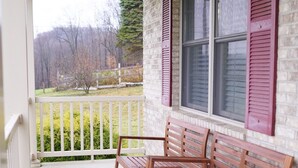 Une véranda avec deux bancs en bois et une table blanche, un mur en briques et une fenêtre avec des volets.
