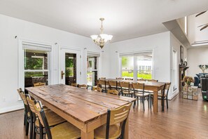 A spacious dining area with a long wooden table and multiple chairs, large windows, and a chandelier.