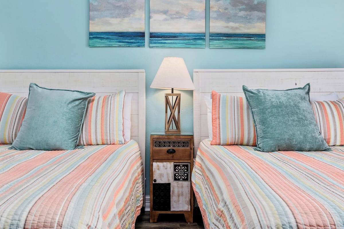 Una habitación con dos camas, cada una con ropa de cama a rayas y almohadas turquesa, una mesita de noche con una lámpara, y tres cuadros del océano en la pared.