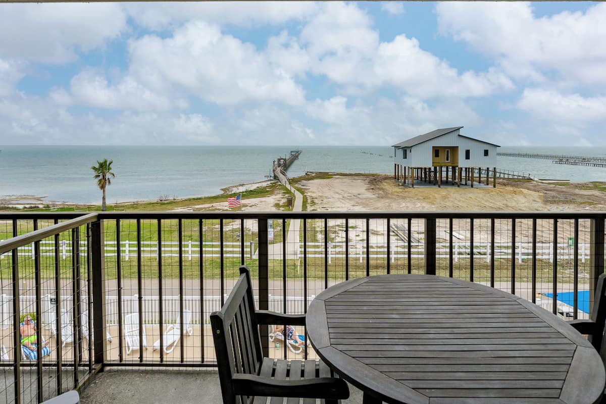 Un balcón con vista a la playa, una mesa de madera y una casa pequeña con un muelle al fondo.