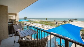 Un balcón con vista a la playa y al océano, un área de piscina con sillas de descanso y una mesa con sillas.
