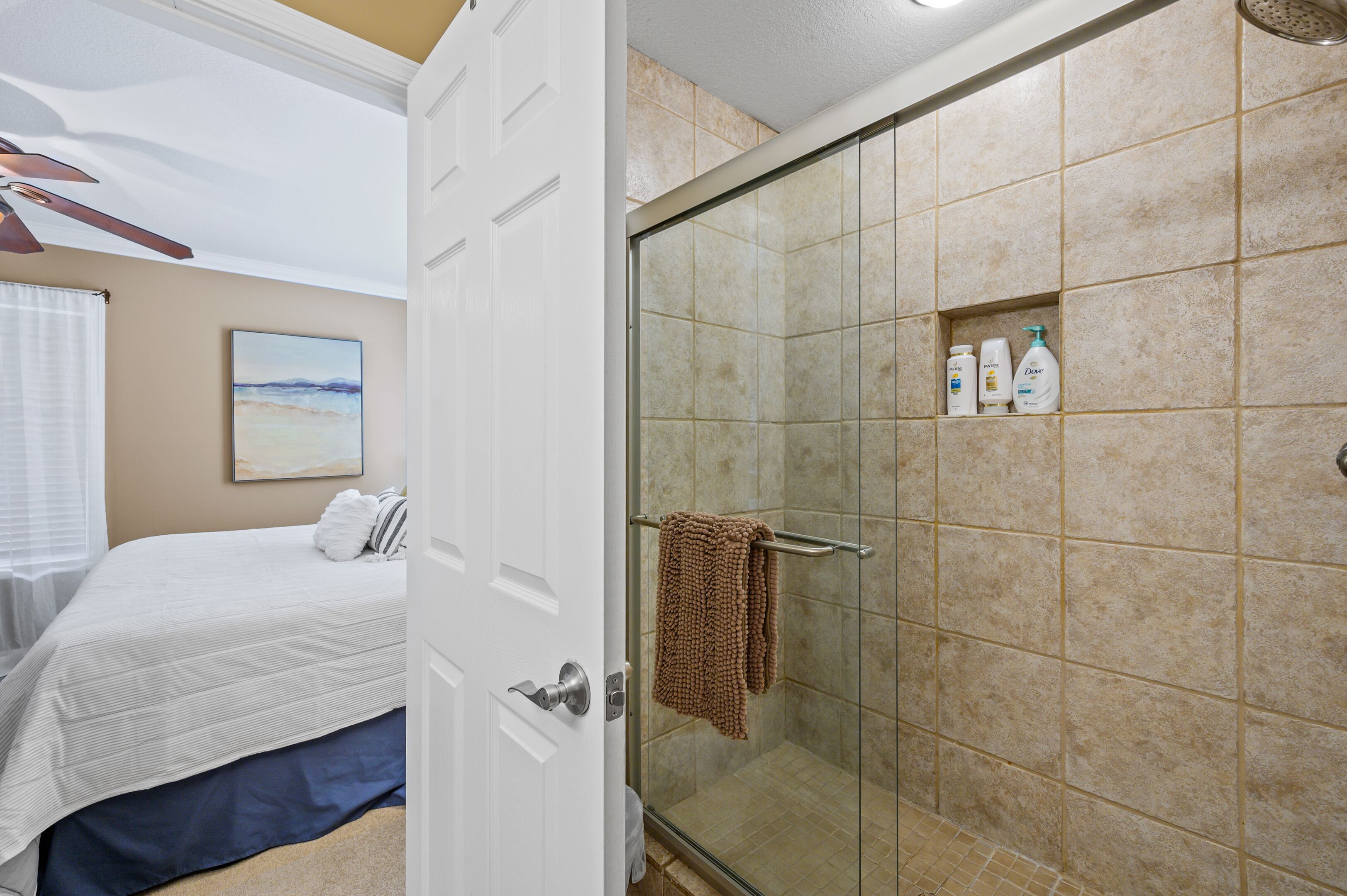 A hotel room with a bed, a shower area with beige tiles, and a door leading to another room.