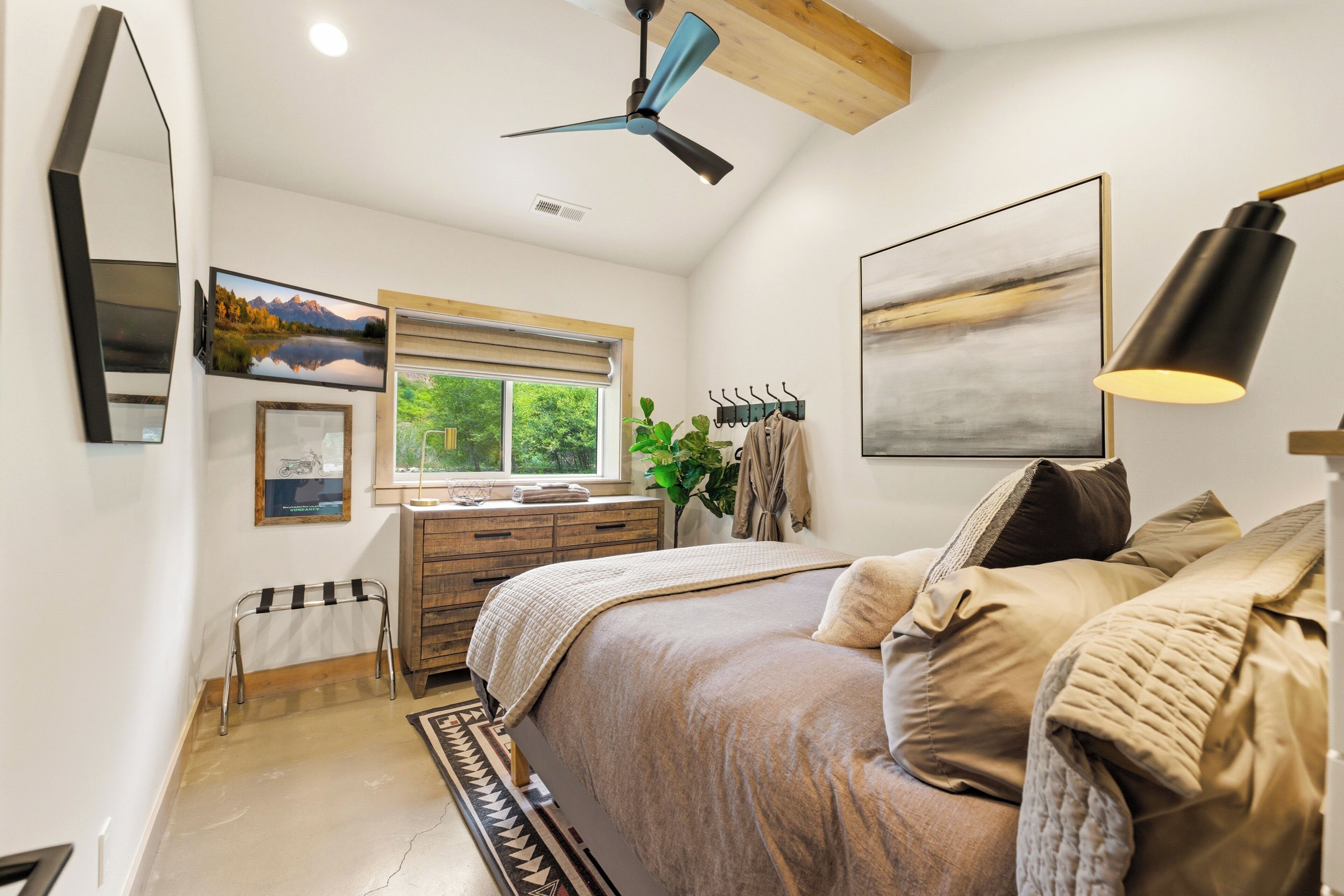 A bedroom with a bed, a dresser, a mirror, a ceiling fan, and a window with a view of greenery.