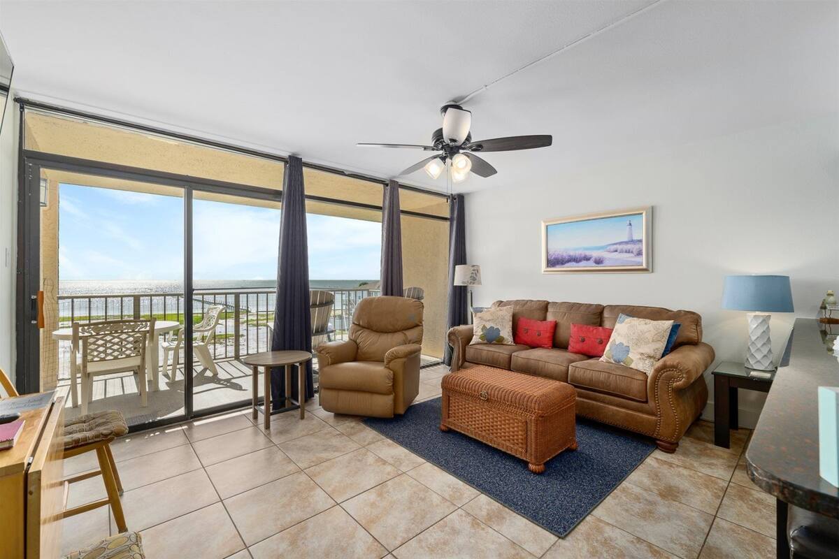 A spacious living area with a balcony, a ceiling fan, a sofa set, a wicker ottoman, and a view of the ocean.