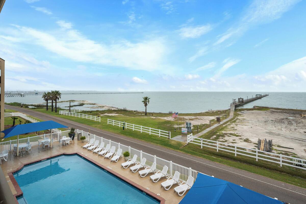 A pool with lounge chairs, a view of the beach, and a pier in the distance.