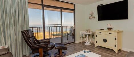 A living room with a view of the beach, a ceiling fan, a TV, and a small table with magazines.