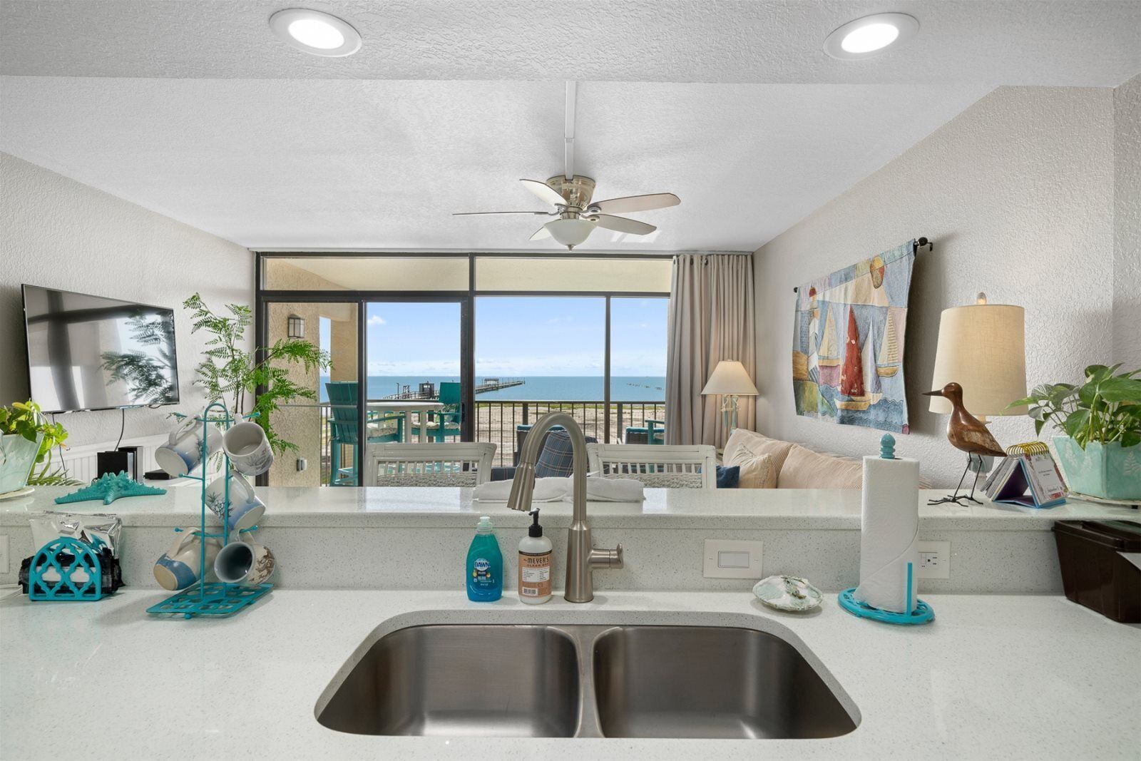 A modern hotel room with a balcony, a large window, a ceiling fan, a TV, and a kitchen area with a sink and countertop.