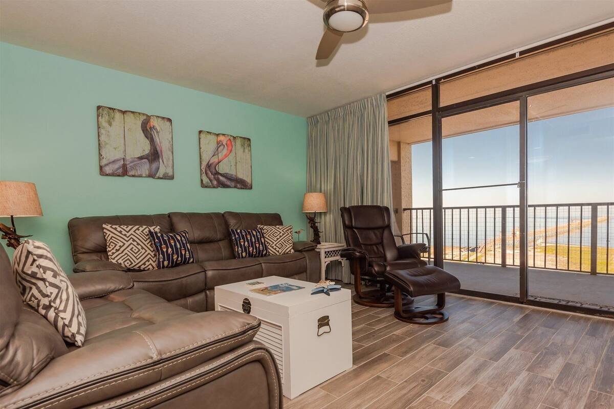 A living room with a brown leather sofa, patterned cushions, a white coffee table, and a reclining chair. There are two paintings on the wall, a ceiling fan, and a large sliding glass door leading to a balcony with a view of the beach.