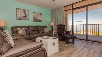 Un salón con un sofá de cuero marrón, cojines estampados, una mesa de centro blanca y un sillón reclinable. Hay dos cuadros en la pared, un ventilador de techo y un amplio ventanal corredero que da a un balcón con vistas a la playa.