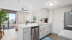 A modern kitchen with white cabinetry, a dishwasher, and a refrigerator. There is a dining area with a table and chairs, and a balcony with a view. - Laguna Reef 2 by Miss Kitty’s (Rockport)