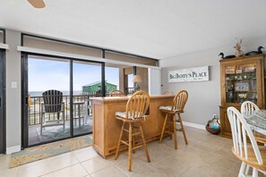 A dining area with a wooden bar, two wooden chairs, a white table, and a cabinet with glass doors. - Laguna Reef 2 by Miss Kitty’s (Rockport)