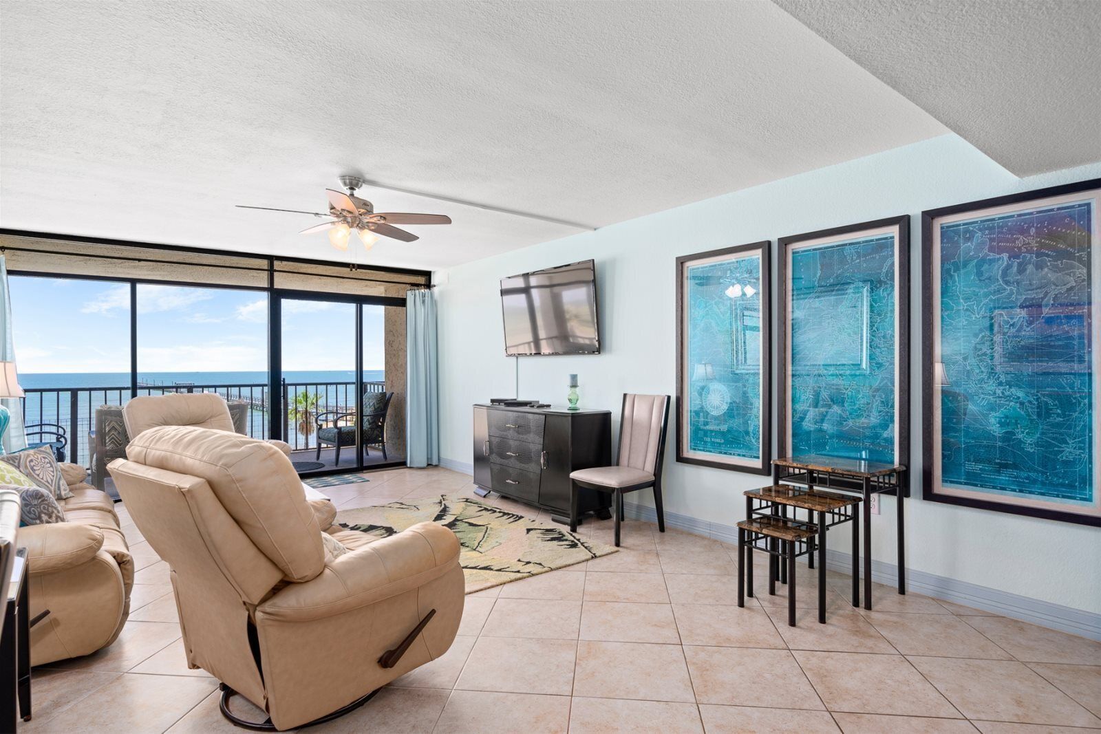 A spacious living area with a large sliding glass door leading to a balcony with a view of the ocean. There is a beige recliner, a small table with a lamp, and framed maps on the wall.