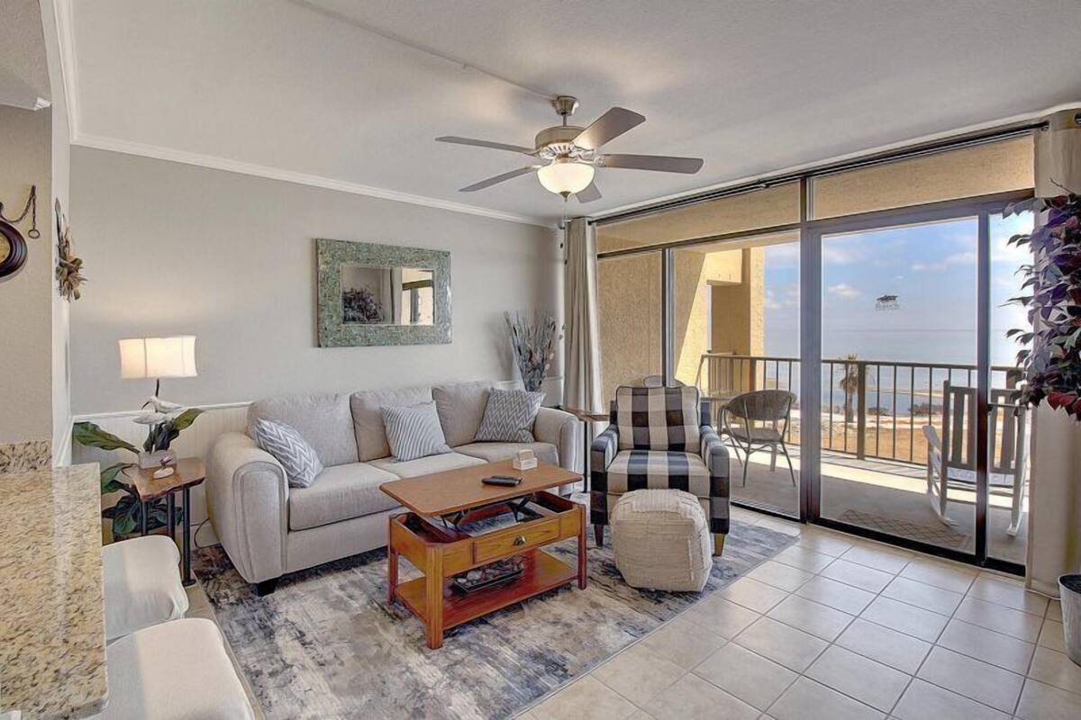 A living room with a sofa, coffee table, and a chair. There is a ceiling fan, a window with a view of the beach, and a balcony with outdoor furniture.