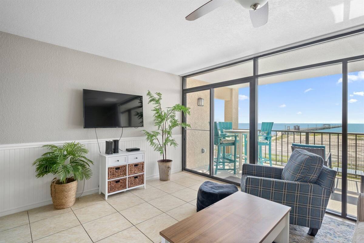 A living room with a TV, a plant, a coffee table, and a balcony with a view of the beach.