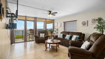A spacious living room with a large sofa, armchairs, and a coffee table. There is a balcony with a view of the water.