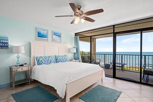 A bedroom with a bed, a ceiling fan, a nightstand, a lamp, a view of the ocean, and a balcony with outdoor seating.