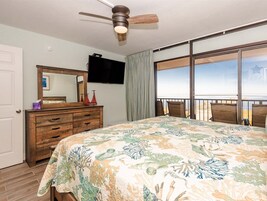 Un dormitorio con cama, cómoda, televisor y balcón con vista a la playa.
