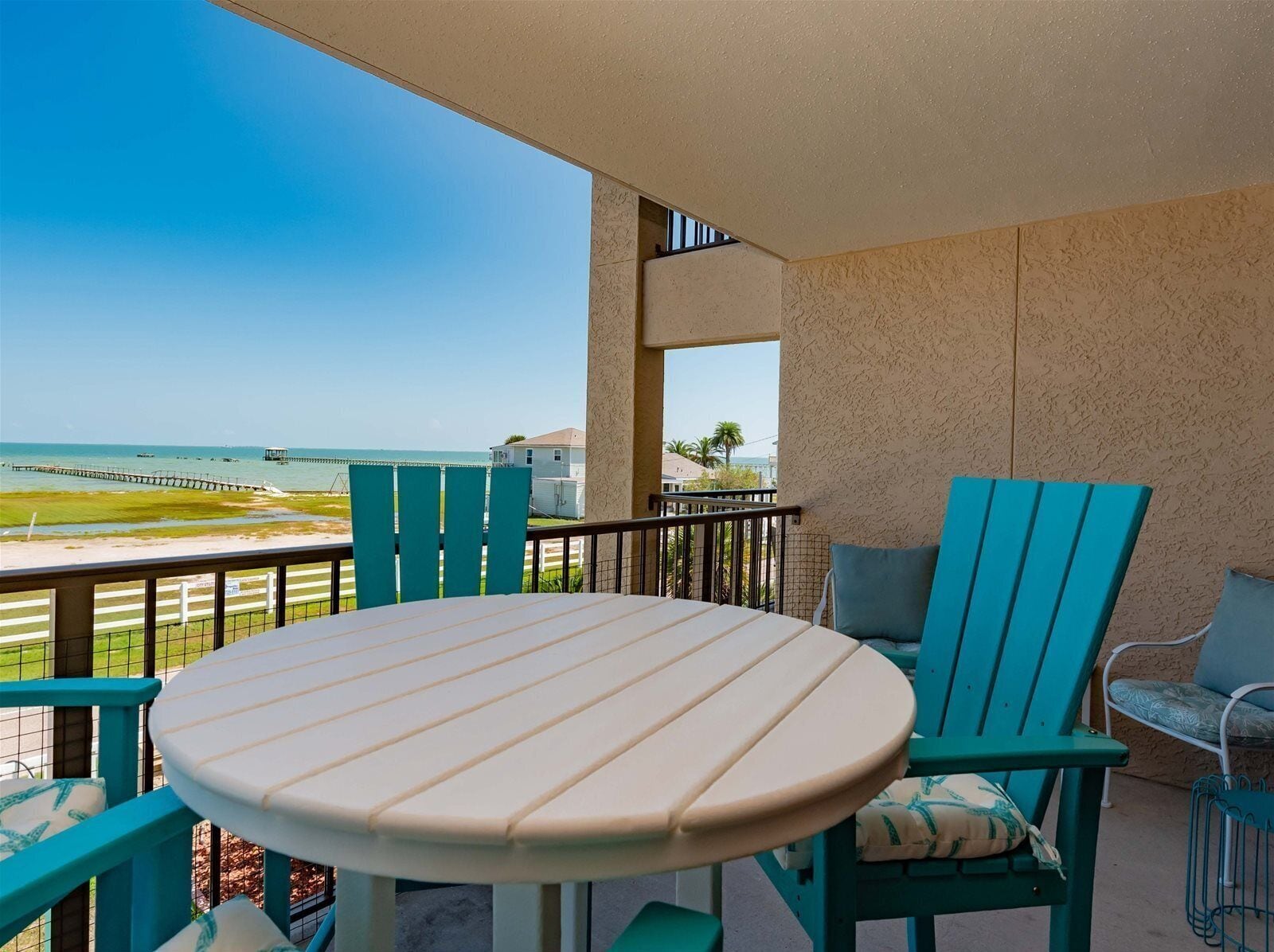 A balcony with a round table and two chairs, overlooking a beach with a pier.