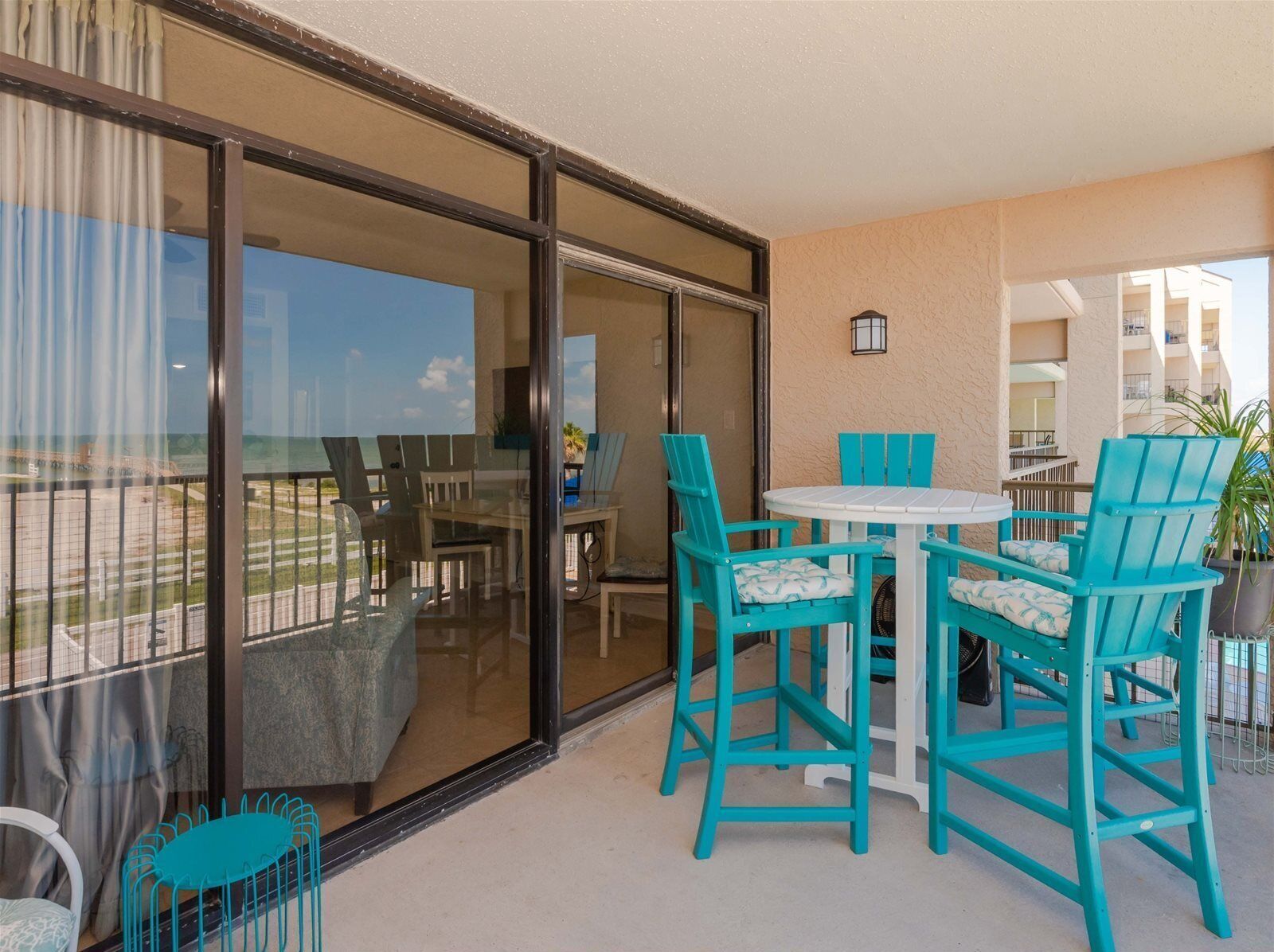 A balcony with turquoise chairs and a round table, overlooking a beach and buildings.