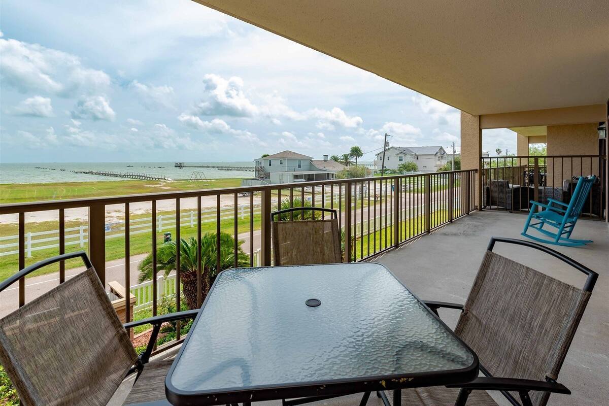 A balcony with a table and chairs, overlooking a beach and houses.