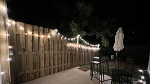 Un patio con cerco de madera, luces colgantes, una mesa con sillas y un paraguas blanco.