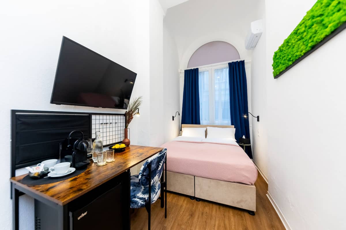 A compact hotel room with a bed, a desk with a coffee maker, a TV, and a small seating area.