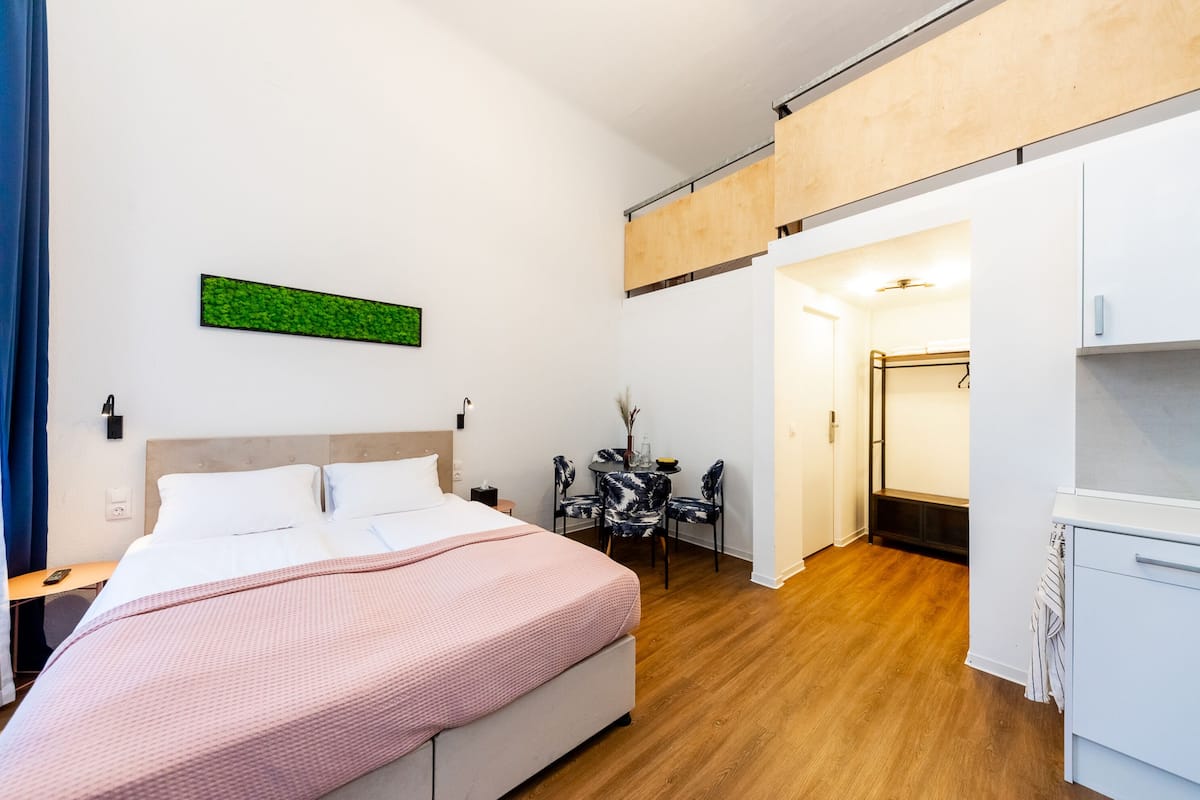 A modern hotel room with a bed, desk, and chair, a small table with a vase, and a green wall decoration.